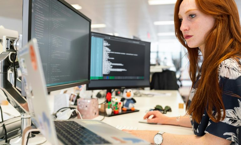 A focused female software engineer coding on dual monitors in a modern office.