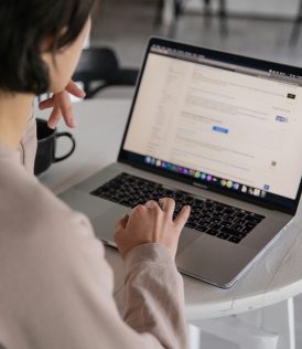 Back view of crop anonymous female remote employee typing on portable computer while watching website on screen and sitting at plastic table with cup of hot drink in apartment