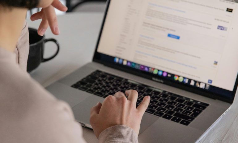 Back view of crop anonymous female remote employee typing on portable computer while watching website on screen and sitting at plastic table with cup of hot drink in apartment