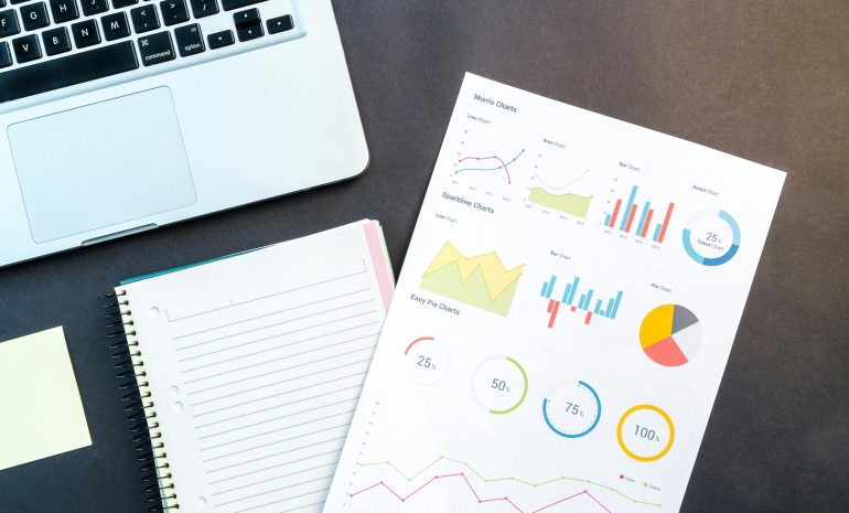 Flatlay of business report with colorful charts, a notebook, and a laptop for data analysis on a desk.